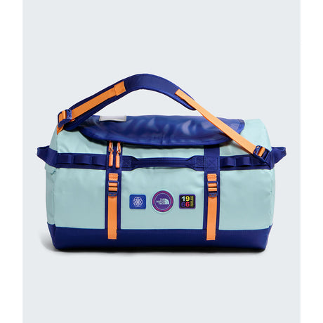 A light blue and navy duffel bag with bright orange straps and accents, featuring multiple brand patches on the front, set against a light gray background. - The North Face Base Camp Duffel Small 50L - Canal Blue/Bold Blue/Papaya