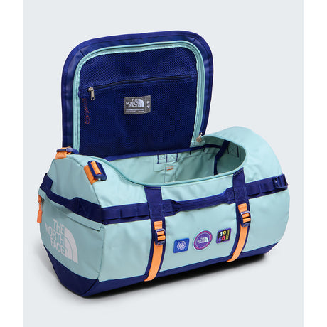 A light blue and navy blue duffel bag by The North Face, shown open to display its interior. The bag features orange accents on the straps and zippers, and various brand patches on the front. - The North Face Base Camp Duffel Small 50L - Canal Blue/Bold Blue/Papaya