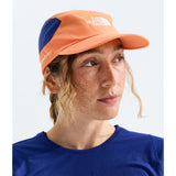 A woman wearing an orange and navy blue The North Face running cap, shown in a close-up profile against a light gray background. - The North Face Summer LIGHTRANGE Run Hat - Papaya