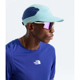 A man in profile wearing a light blue and navy blue The North Face Summit Series cap, white-framed sunglasses with pink mirrored lenses, and a royal blue The North Face athletic shirt against a light gray background. - The North Face Summer LIGHTRANGE Run Hat - Canal Blue