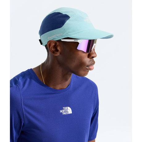 A man in profile wearing a light blue and navy blue The North Face Summit Series cap, white-framed sunglasses with pink mirrored lenses, and a royal blue The North Face athletic shirt against a light gray background. - The North Face Summer LIGHTRANGE Run Hat - Canal Blue