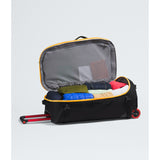 An open black rolling suitcase with yellow trim, shown with its telescoping handle extended and packed with folded clothes inside, on a light gray studio background. - The North Face Base Camp Rolling Thunder 28" (95L) - Summit Gold/TNF Gold-NPF
