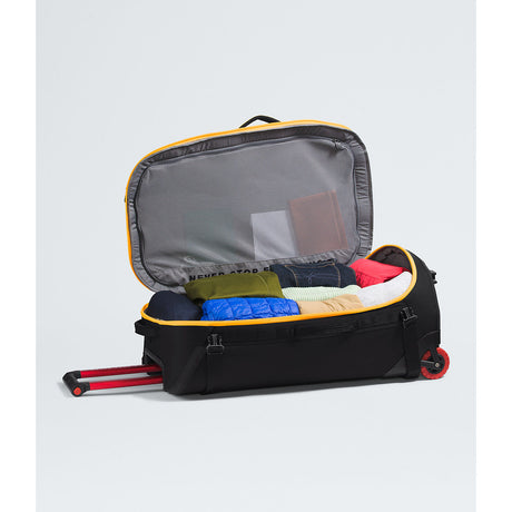 An open black rolling suitcase with yellow trim, shown with its telescoping handle extended and packed with folded clothes inside, on a light gray studio background. - The North Face Base Camp Rolling Thunder 28" (95L) - Summit Gold/TNF Gold-NPF