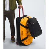 Person holding a yellow suitcase and black backpack against a light gray background - The North Face Base Camp Rolling Thunder 28" (95L) - Summit Gold/TNF Gold-NPF