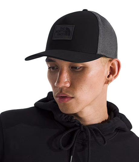 A man wearing a black trucker hat with a black mesh back and a black patch logo for The North Face, paired with a black hoodie against a white background. - The North Face Keep It Patched Trucker - TNF Black/Asphalt Grey