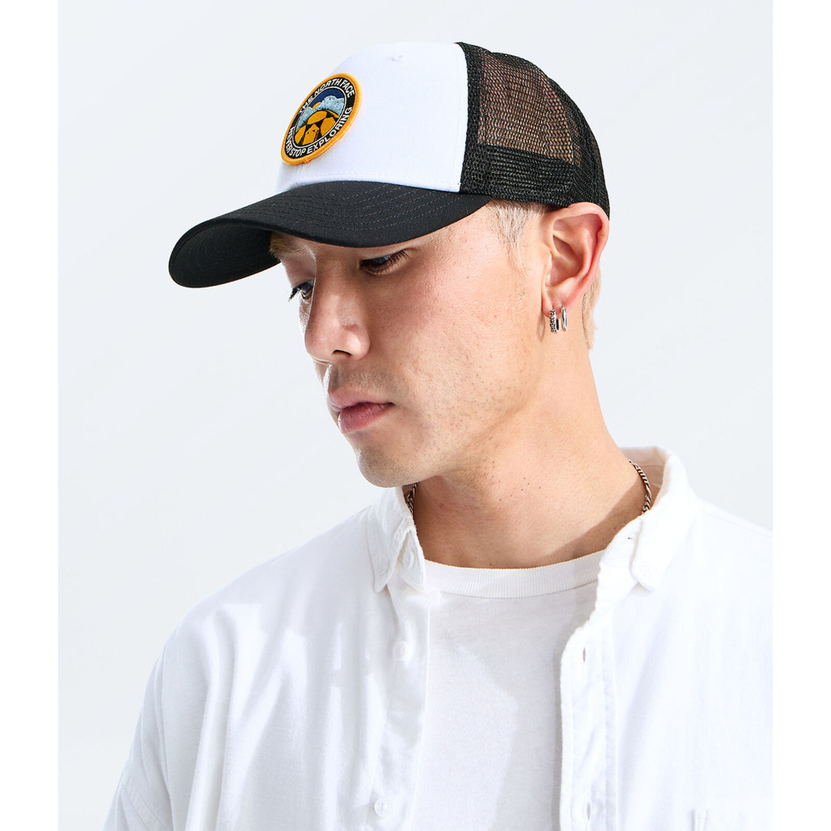 A man wearing a black and white trucker hat with a circular embroidered logo on the front, paired with a white button-down shirt, photographed against a clean white background. - The North Face Graphic Mudder Trucker - TNF White/TNF Black/TNF NSE Dome Patch