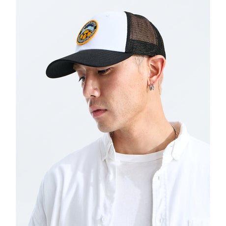 A man wearing a black and white trucker hat with a circular embroidered logo on the front, paired with a white button-down shirt, photographed against a clean white background. - The North Face Graphic Mudder Trucker - TNF White/TNF Black/TNF NSE Dome Patch