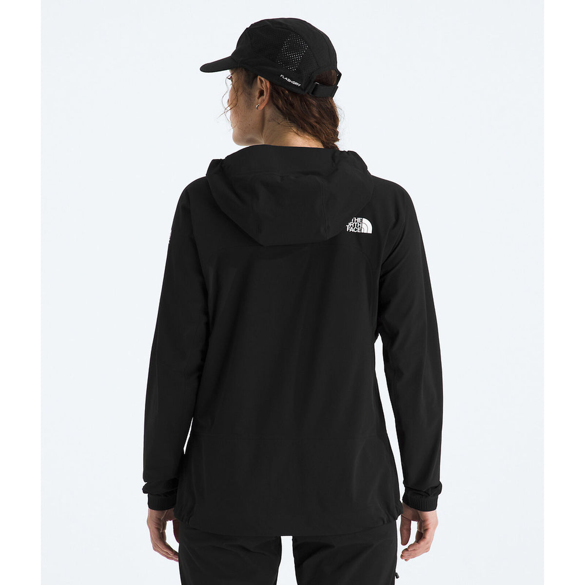 A woman viewed from behind wearing a black hooded jacket with a small white The North Face logo on the back, along with a black cap, against a light gray studio background. - The North Face Summit Series Off Width Jacket (Women's) - TNF Black