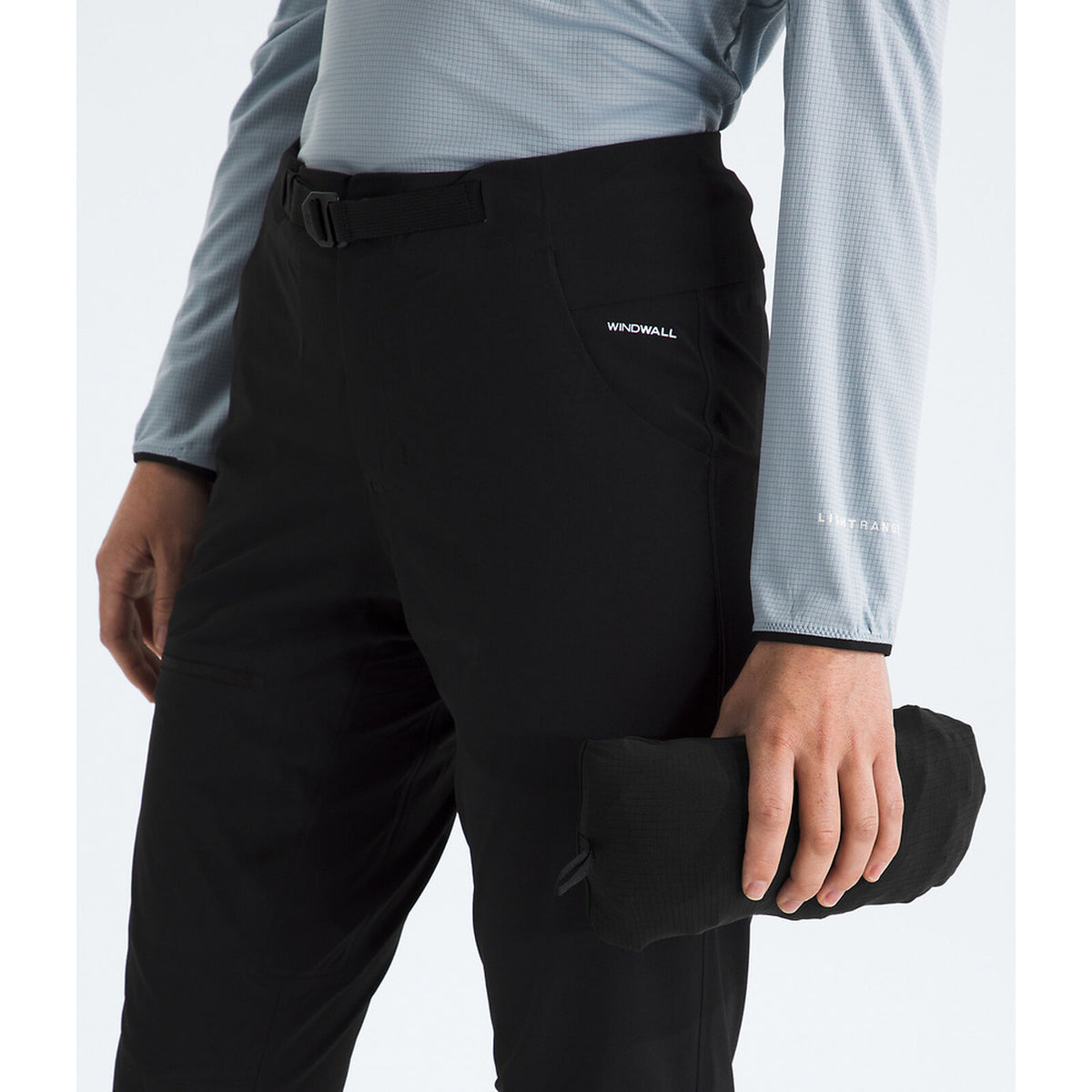 A close-up of a person wearing black Wyonull outdoor pants with a matching black belt, paired with a light blue long-sleeve shirt. The person is holding a rolled-up black garment against a clean white background. - The North Face Summit Series Off Width Jacket (Women's) - TNF Black