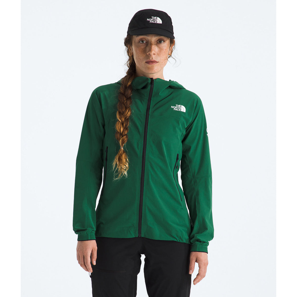 A woman wearing a green The North Face zip-up hooded jacket, a black The North Face cap, and black pants, standing against a light gray studio background. - The North Face Summit Series Off Width Jacket (Women's) - Evergreen