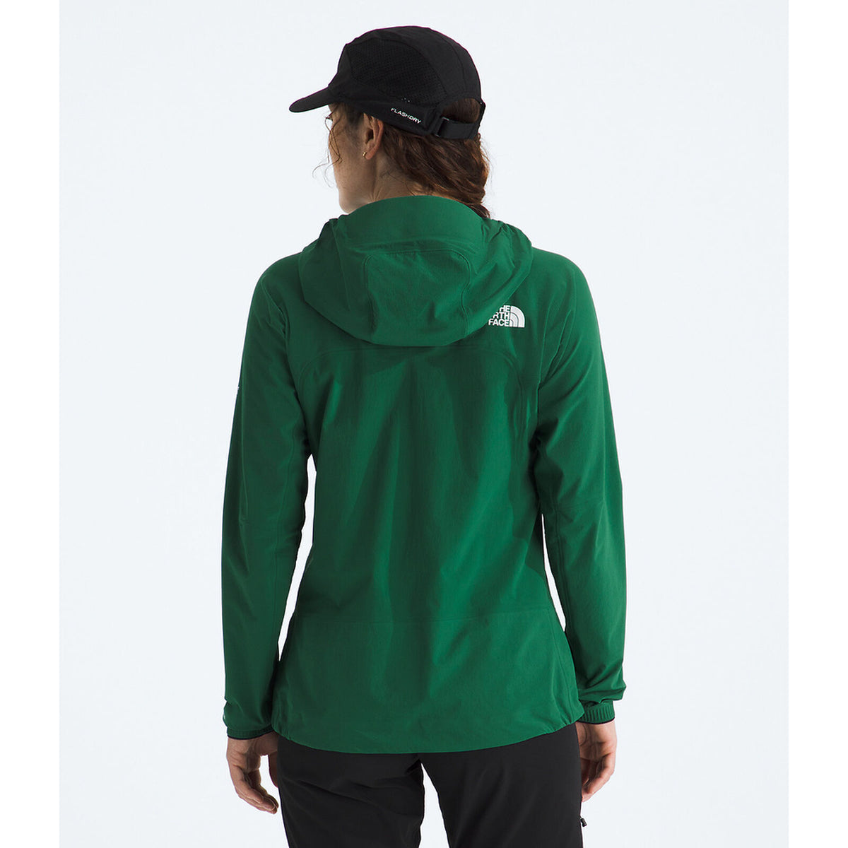 A woman viewed from the back wearing a dark green hooded jacket with a small white The North Face logo on the upper back, a black cap, and black pants, against a plain white background. - The North Face Summit Series Off Width Jacket (Women's) - Evergreen