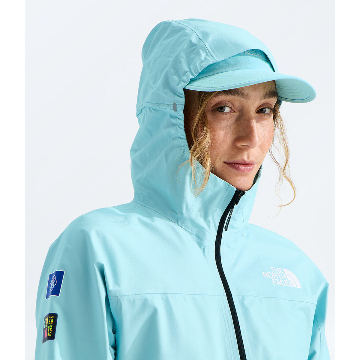 A woman wearing a light blue hooded jacket and matching visor from The North Face, shown against a clean white studio background. - The North Face Summit FUTURELIGHT Superior Jacket-Graphic (Women's) - Canal Blue