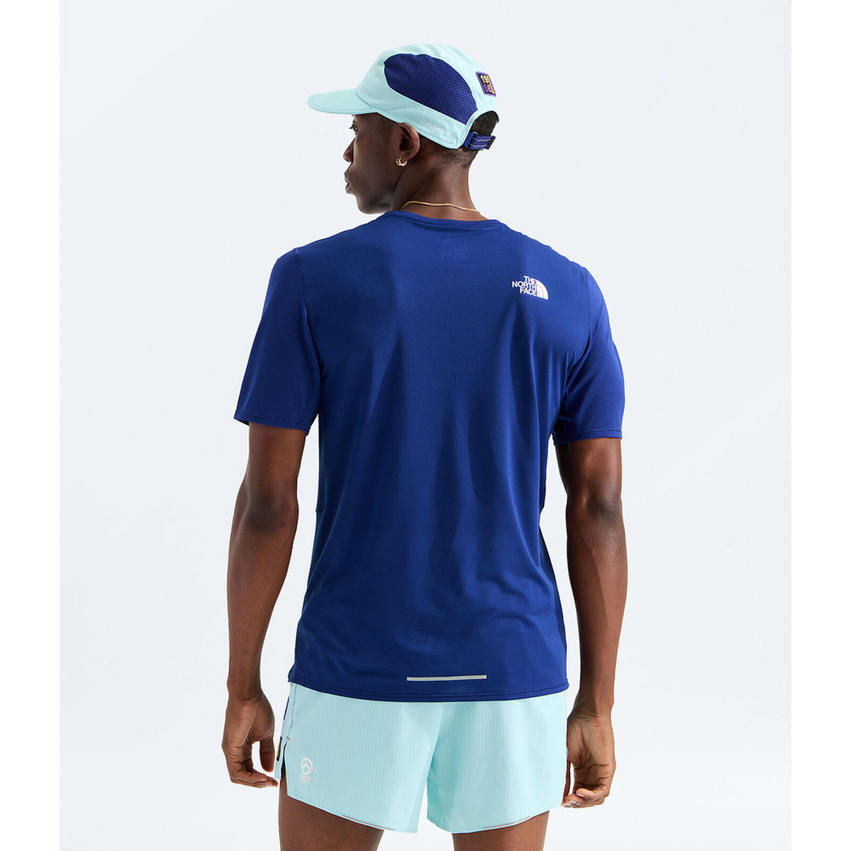 A man wearing a royal blue short-sleeve athletic t-shirt from the back, paired with light blue shorts and a matching light blue and navy cap, against a plain white background. - The North Face Summit High Trail Short Sleeve-Graphic (Men's) - Bold Blue