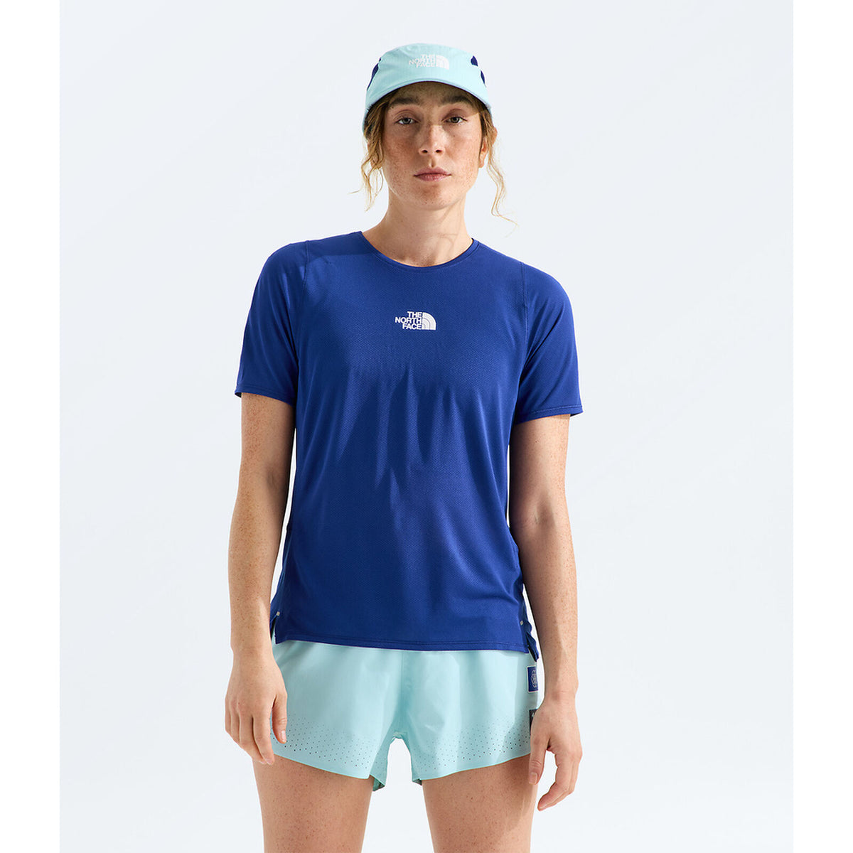 A woman modeling a royal blue short-sleeve athletic t-shirt by The North Face, paired with light blue shorts and a matching light blue cap, against a plain light gray studio background. - The North Face Summit High Trail Short Sleeve-Graphic (Women's) - Bold Blue