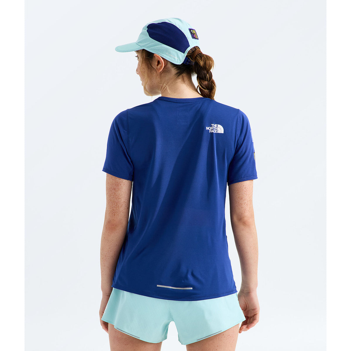 A woman shown from the back wearing a royal blue short-sleeve athletic t-shirt by The North Face, paired with mint green shorts and a matching two-tone cap, against a plain light gray background. - The North Face Summit High Trail Short Sleeve-Graphic (Women's) - Bold Blue