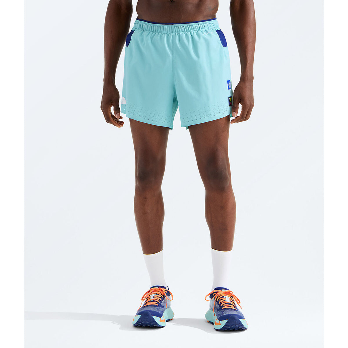 A model from the waist down wearing light blue athletic shorts with dark blue side accents, white crew socks, and blue running shoes with orange laces, set against a plain white studio background. - The North Face Summit Series Pacesetter 5" Run Shorts-Graphic (Men's) - Canal Blue