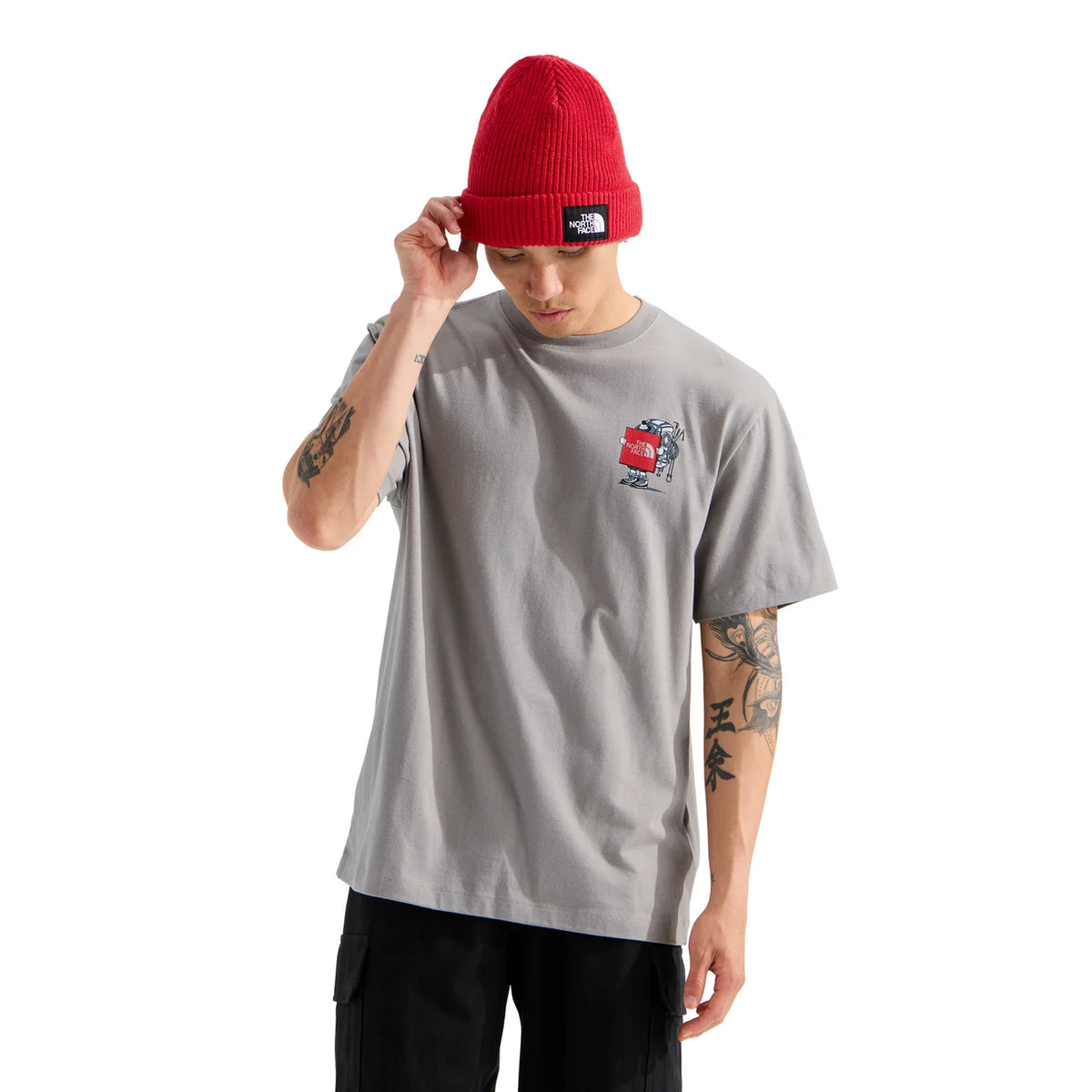 A man wearing a light gray crewneck t-shirt with a small red and black graphic on the left chest, paired with a red knit beanie and black pants, against a plain white background. - The North Face Gear Up Get Out Relaxed Short-Sleeve Tee-Graphic (Unisex) - Stone Slab