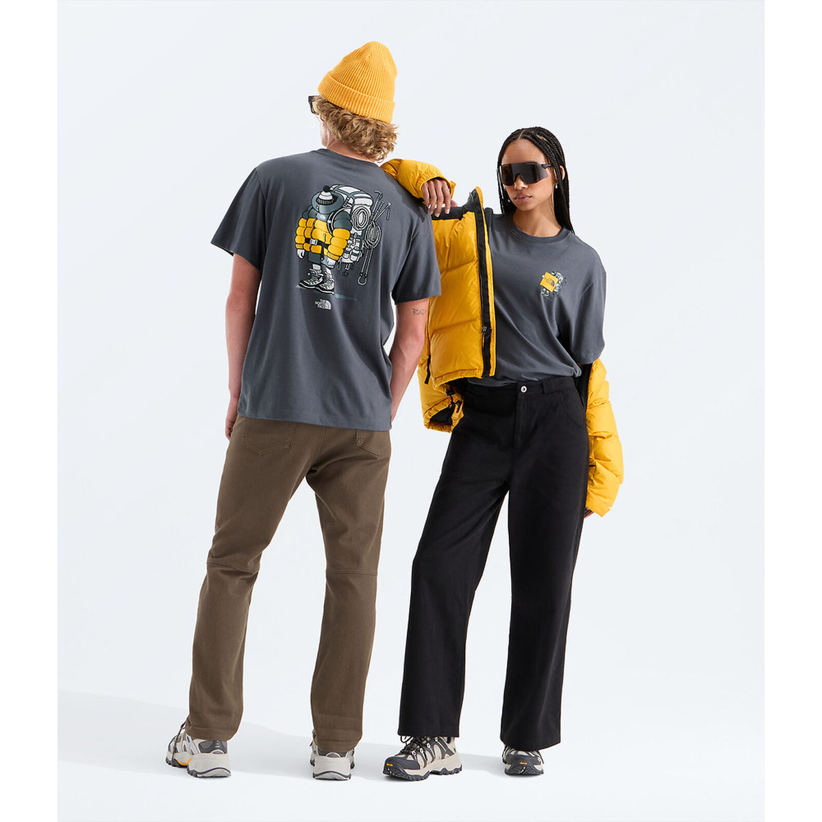 Two models wearing dark gray t-shirts with graphic prints, styled with brown and black pants, a yellow puffer jacket, and a yellow beanie, set against a plain white studio background. - The North Face Gear Up Get Out Relaxed Short-Sleeve Tee-Graphic (Unisex) - Anthracite Grey