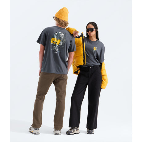 Two models wearing dark gray t-shirts with graphic prints, styled with brown and black pants, a yellow puffer jacket, and a yellow beanie, set against a plain white studio background. - The North Face Gear Up Get Out Relaxed Short-Sleeve Tee-Graphic (Unisex) - Anthracite Grey