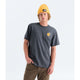 A man wearing a dark charcoal gray t-shirt with a small yellow graphic on the left chest, paired with a yellow beanie and brown pants, standing against a light gray studio background. - The North Face Gear Up Get Out Relaxed Short-Sleeve Tee-Graphic (Unisex) - Anthracite Grey