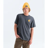 A man wearing a dark charcoal gray t-shirt with a small yellow graphic on the left chest, paired with a yellow beanie and brown pants, standing against a light gray studio background. - The North Face Gear Up Get Out Relaxed Short-Sleeve Tee-Graphic (Unisex) - Anthracite Grey