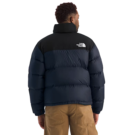 A man viewed from the back wearing a puffer jacket with a black upper section and a dark navy lower section, paired with tan cargo pants against a plain white background. - The North Face 1996 Retro Nuptse Jacket (Men's) - Summit Navy/TNF Black