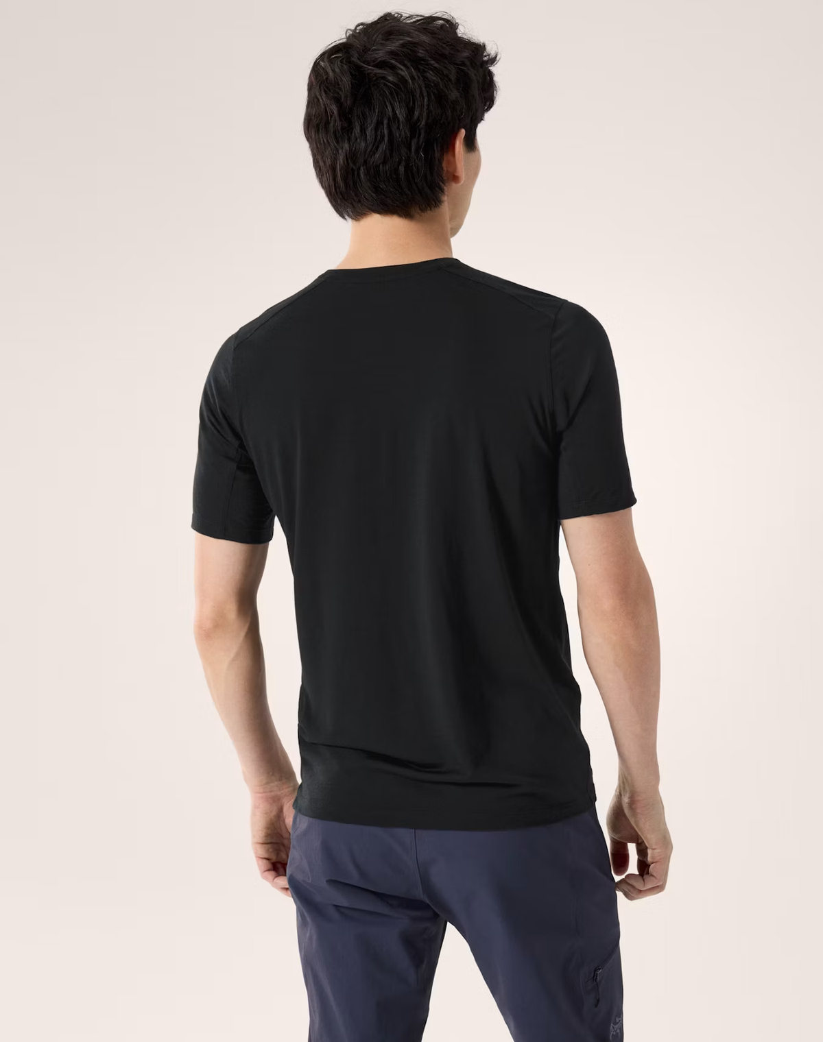 A man viewed from the back wearing a black short-sleeve crewneck t-shirt and navy blue pants, set against a plain light beige studio background. - Arc'teryx Merino Wool Shirt Short Sleeve (Men's) - Black