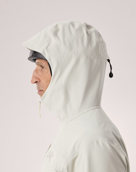 Side profile of a man wearing a light off-white hooded rain jacket, showing the hood drawstring and face shield attachment, against a solid pale pink background. - Arc'teryx Gamma MX Hoody (Men's) - Sea Salt