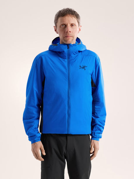 A man wearing a bright blue hooded Arc'teryx jacket and black pants, standing against a plain light beige studio background. - Arc'teryx Atom Hoody (Men's) - Fluidity/Black Sapphire