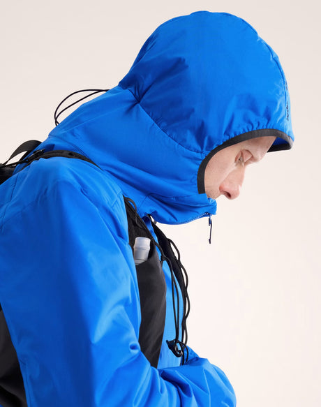 A close-up side profile of a person wearing a bright blue hooded rain jacket with black trim and drawstrings, against a plain light background. - Arc'teryx Atom Hoody (Men's) - Fluidity/Black Sapphire