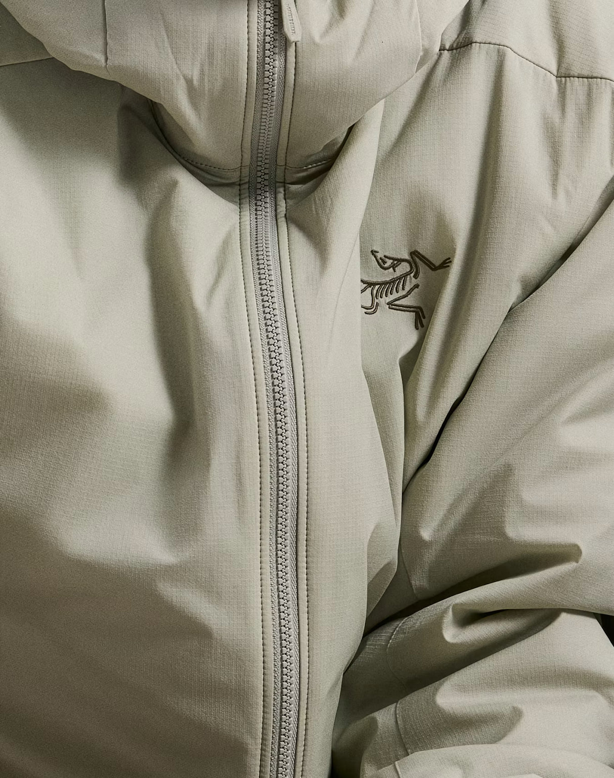 Close-up of a light beige hooded jacket with a central zipper and the Arc'teryx bird logo embroidered on the chest. - Arc'teryx Atom Hoody (Men's) - Habitat