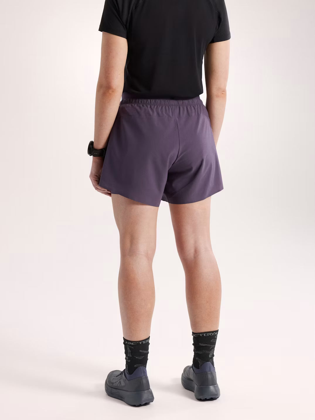 A person shown from the back wearing dark purple athletic shorts, a black t-shirt, black socks, and black shoes against a plain light background. - Arc'teryx Norvan Shorts 5" 2-in-1 (Women's) - Moondrop