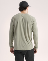 A man viewed from behind wearing a sage green long-sleeve athletic shirt, black pants, and a black baseball cap against a light beige studio background. - Arc'teryx Cormac Crew Neck Shirt Long Sleeve (Men's) - Habitat