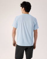 A man viewed from behind wearing a light blue short-sleeve athletic t-shirt and black shorts, standing against a plain light beige studio background. - Arc'teryx Cormac Crew Neck Shirt Short Sleeve (Men's) - Alpine Blue