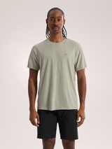 A man wearing a light sage green short-sleeve t-shirt with a small Arc'teryx logo on the chest, paired with black athletic shorts, standing against a light gray studio background. - Arc'teryx Cormac Crew Neck Shirt Short Sleeve (Men's) - Habitat