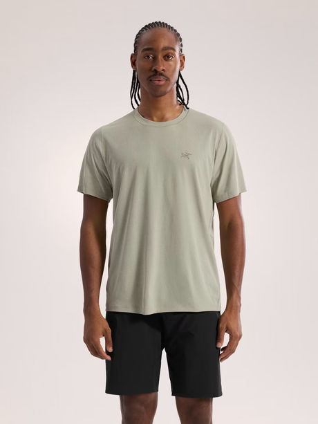 A man wearing a light sage green short-sleeve t-shirt with a small Arc'teryx logo on the chest, paired with black athletic shorts, standing against a light gray studio background. - Arc'teryx Cormac Crew Neck Shirt Short Sleeve (Men's) - Habitat
