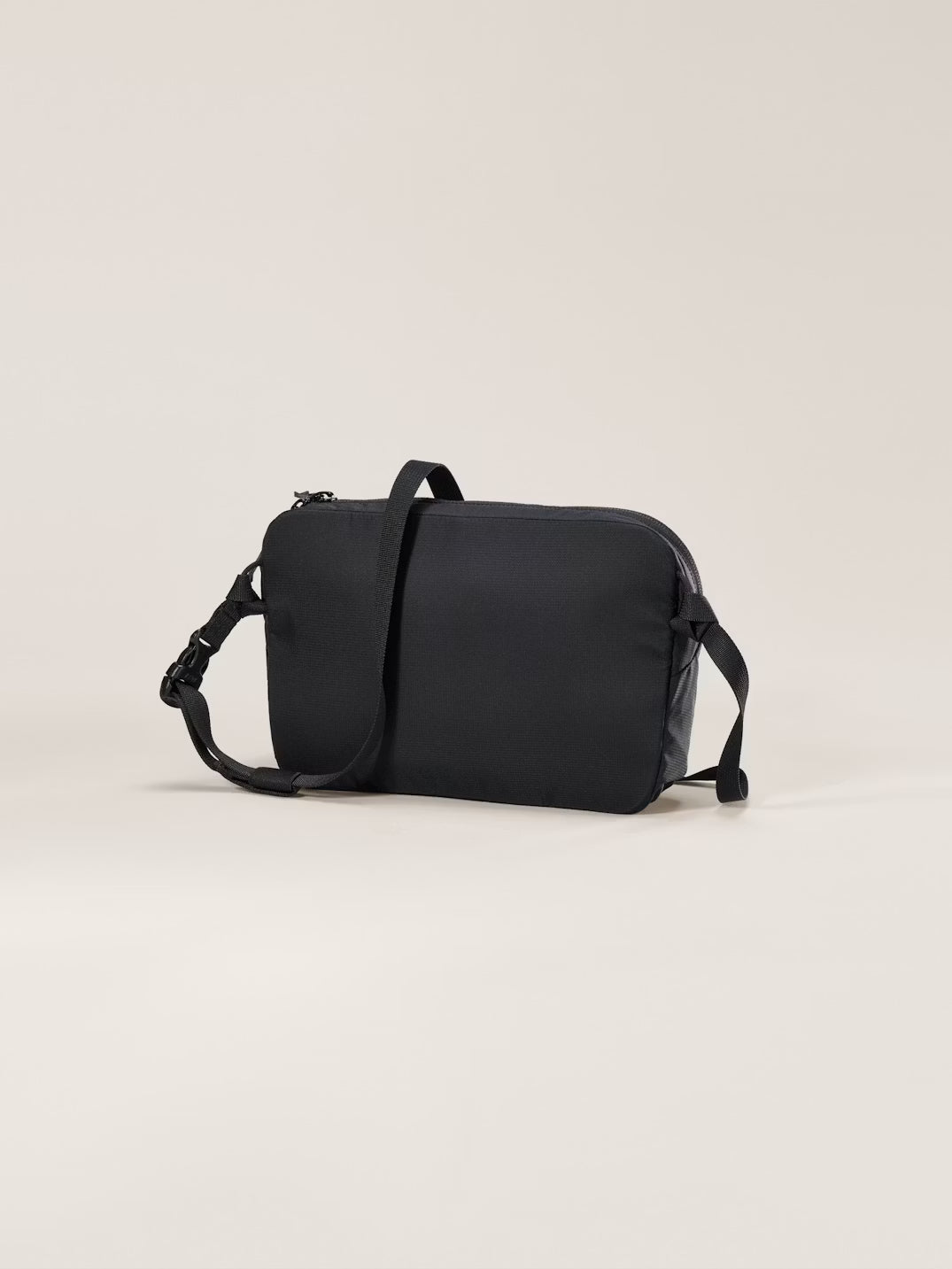 A black rectangular messenger bag with a top handle, adjustable shoulder strap, and zippered closure, photographed on a clean, solid, light beige background. - Arc'teryx Heliad Crossbody Bag - Black