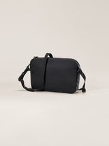 A black rectangular messenger bag with a top handle, adjustable shoulder strap, and zippered closure, photographed on a clean, solid, light beige background. - Arc'teryx Heliad Crossbody Bag - Black