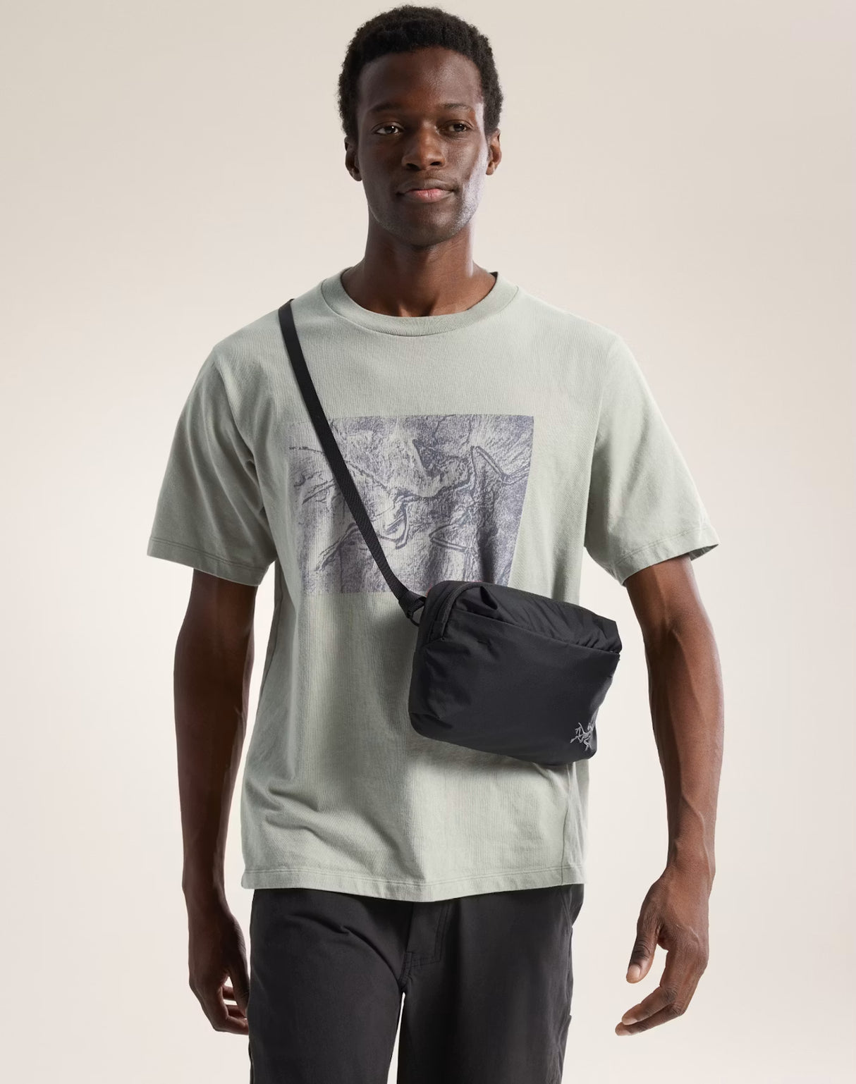 Man wearing a light green t-shirt with a graphic design and carrying a black crossbody bag on a plain background - Arc'teryx Heliad Crossbody Bag - Black