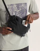 A close-up of a person wearing a black sling bag across their chest, with one hand reaching into the front pocket to place or retrieve a white smartphone. The person is wearing a light sage green graphic t-shirt. - Arc'teryx Heliad Crossbody Bag - Black