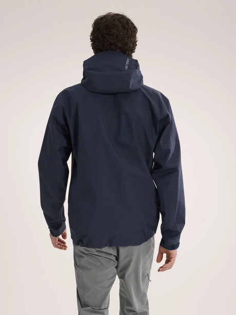 A man viewed from behind wearing a dark navy hooded jacket and light gray pants, standing against a plain light beige studio background. - Arc'teryx Beta Jacket (Men's) - Black Sapphire