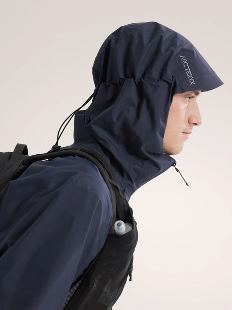 A man in profile wearing a dark navy hooded rain jacket and a black backpack, shown against a plain light background. The Arc'teryx logo is visible on the hood. - Arc'teryx Beta Jacket (Men's) - Black Sapphire