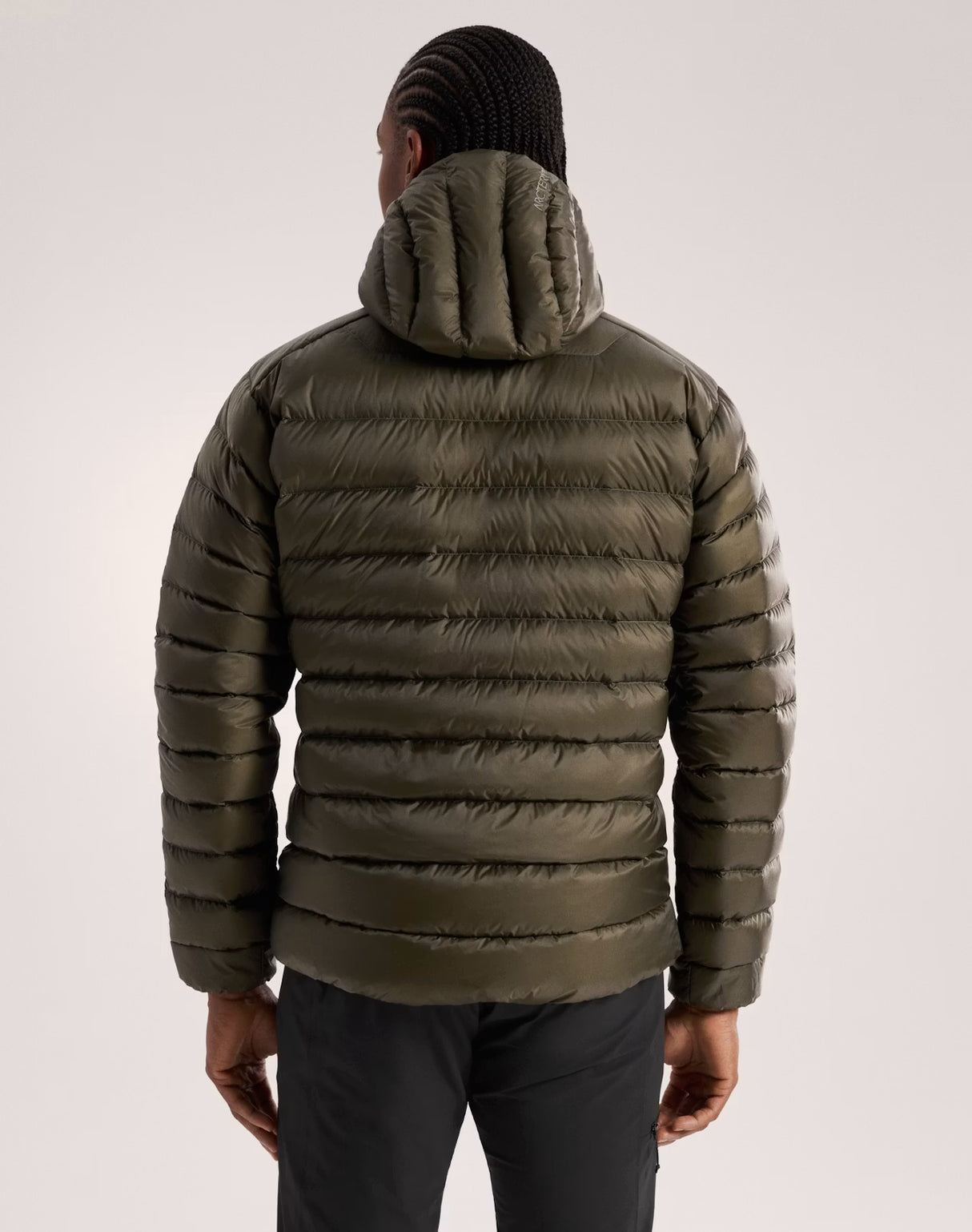 A man viewed from behind wearing an olive green hooded puffer jacket with horizontal quilting, paired with dark trousers and a black knit beanie, against a light gray studio background. - Arc'teryx Cerium Hoody (Men's) - Tatsu II