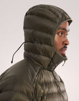 A close-up side profile of a person wearing an olive green Arc'teryx puffer jacket with the hood up, against a plain light gray studio background. - Arc'teryx Cerium Hoody (Men's) - Tatsu II