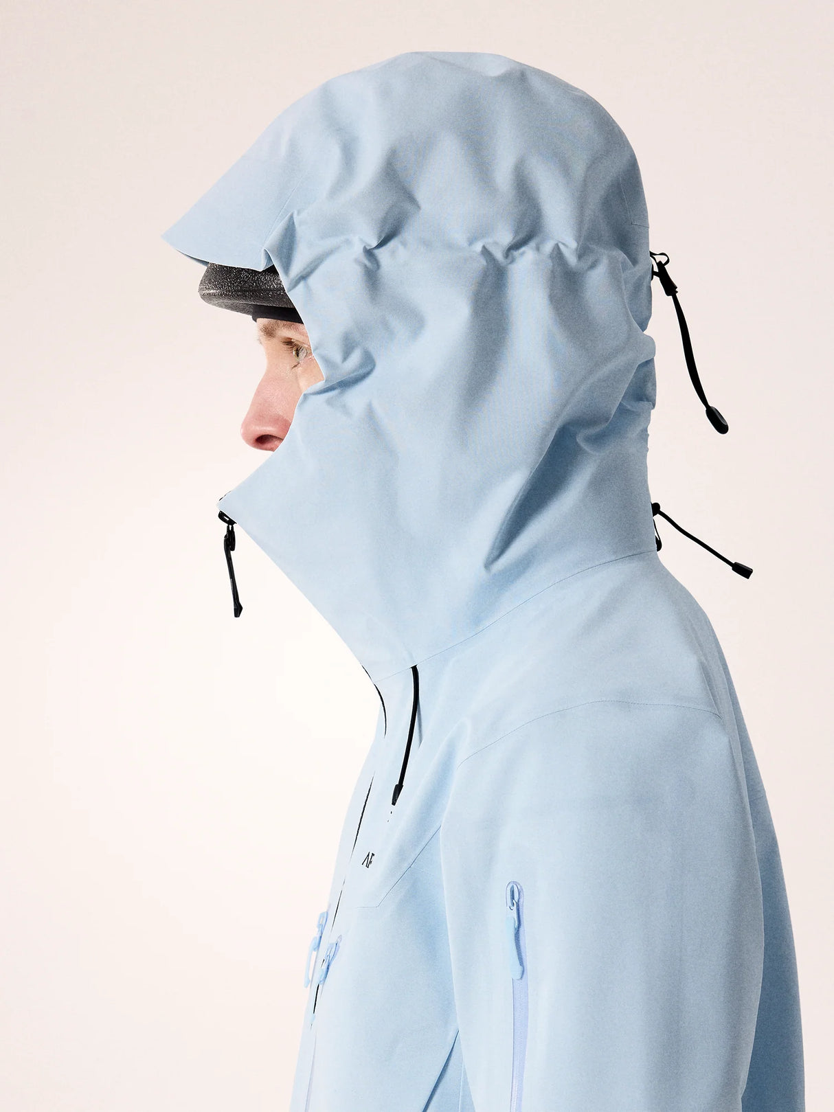 A person shown in side profile wearing a light blue hooded jacket against a plain light beige studio background. - Arc'teryx Alpha SV Jacket (Women's) - Alpine Blue/Black