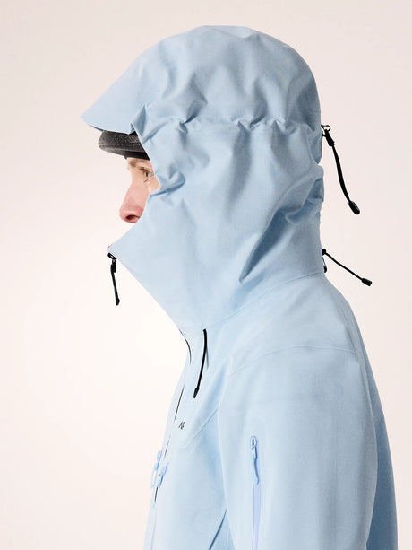 A person shown in side profile wearing a light blue hooded jacket against a plain light beige studio background. - Arc'teryx Alpha SV Jacket (Women's) - Alpine Blue/Black