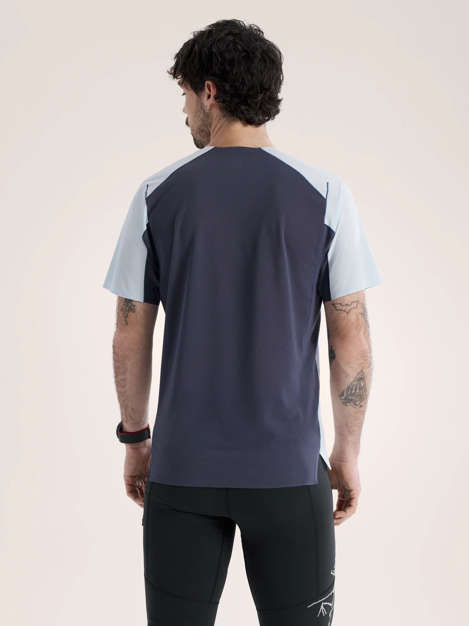 A man viewed from the back wearing a short-sleeve athletic shirt with a dark navy body and light blue shoulder panels, paired with black athletic pants, against a plain light beige studio background. - Arc'teryx Norvan Downword Logo Shirt Short Sleeve (Men's) - Alpine Blue/Dark Stratus