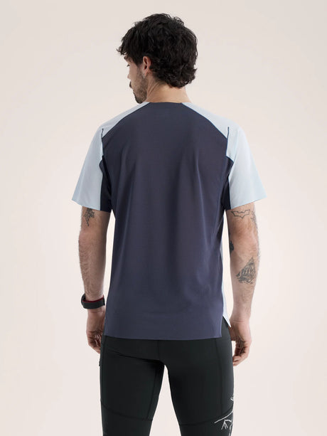 A man viewed from the back wearing a short-sleeve athletic shirt with a dark navy body and light blue shoulder panels, paired with black athletic pants, against a plain light beige studio background. - Arc'teryx Norvan Downword Logo Shirt Short Sleeve (Men's) - Alpine Blue/Dark Stratus