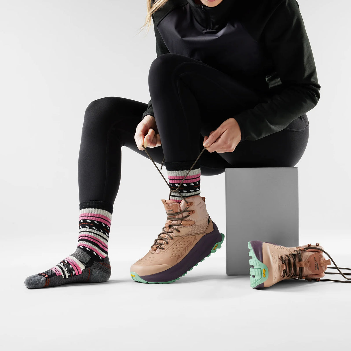 Person sitting on a block wearing black leggings, black jacket, and tan hiking boots with colorful socks. - Smartwool Hike Margarita Light Cushion Crew Socks (Women's) - Black