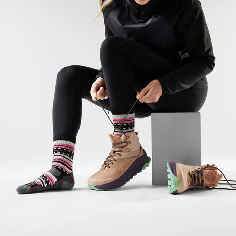 Person sitting on a block wearing black leggings, black jacket, and tan hiking boots with colorful socks. - Smartwool Hike Margarita Light Cushion Crew Socks (Women's) - Black